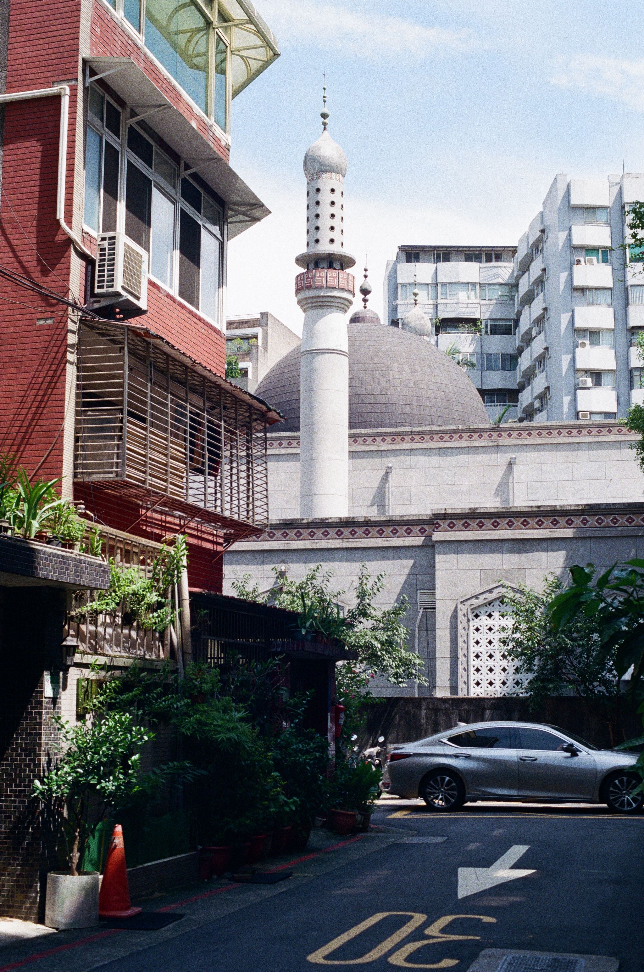 A street scene in a small alley. The frame is dominated by a mosque building in the background. A car drives through the frame in front of the mosque, while a residential apartment building is lit by the midday sun on the left of the shot.