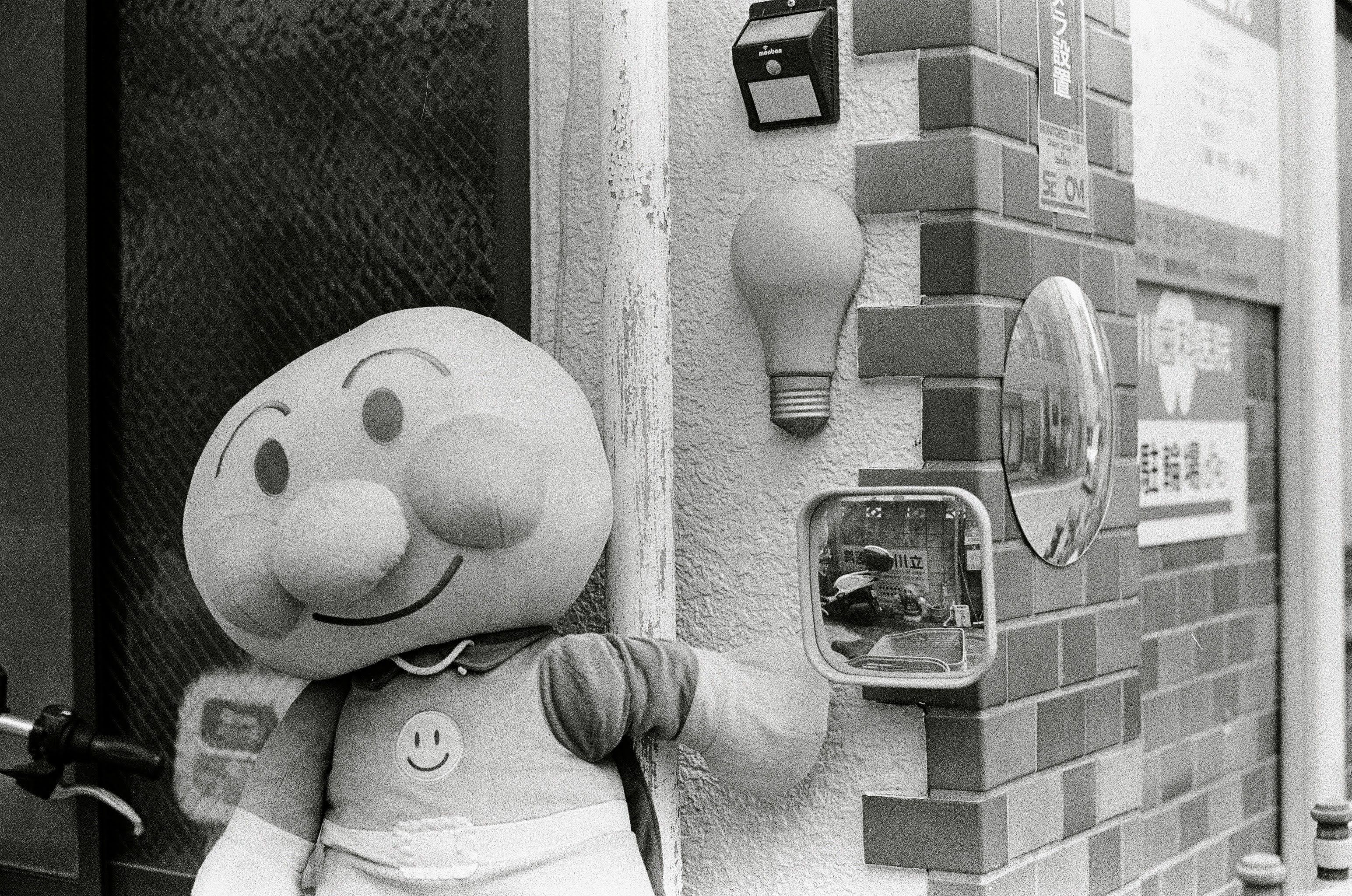 A large Anpanman doll sits outside a house in Tokyo. Next to it is a small mirror which has another smaller Anpanman character reflected in it.